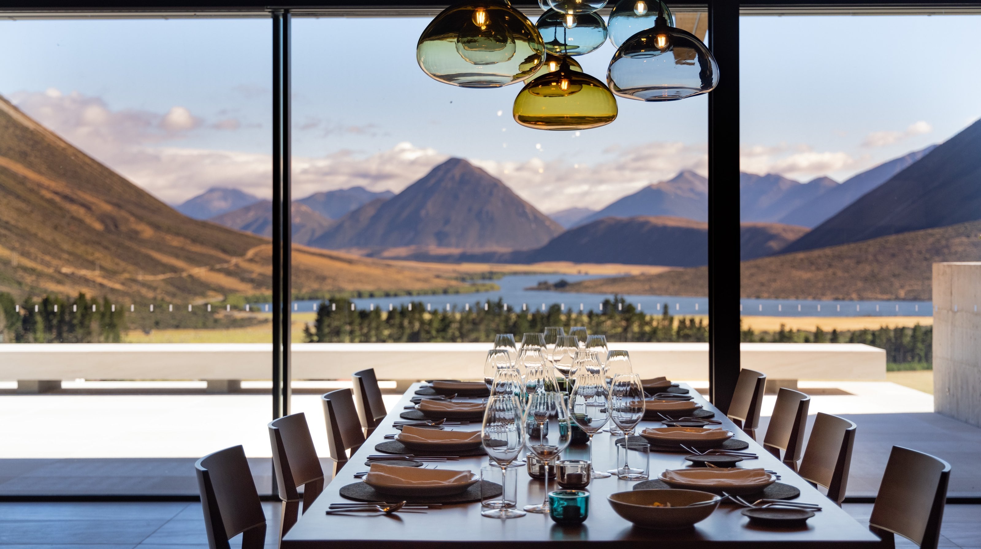 A long dining table set beneath coloured glass pendants at Flockhill, mountains and a lake through the window