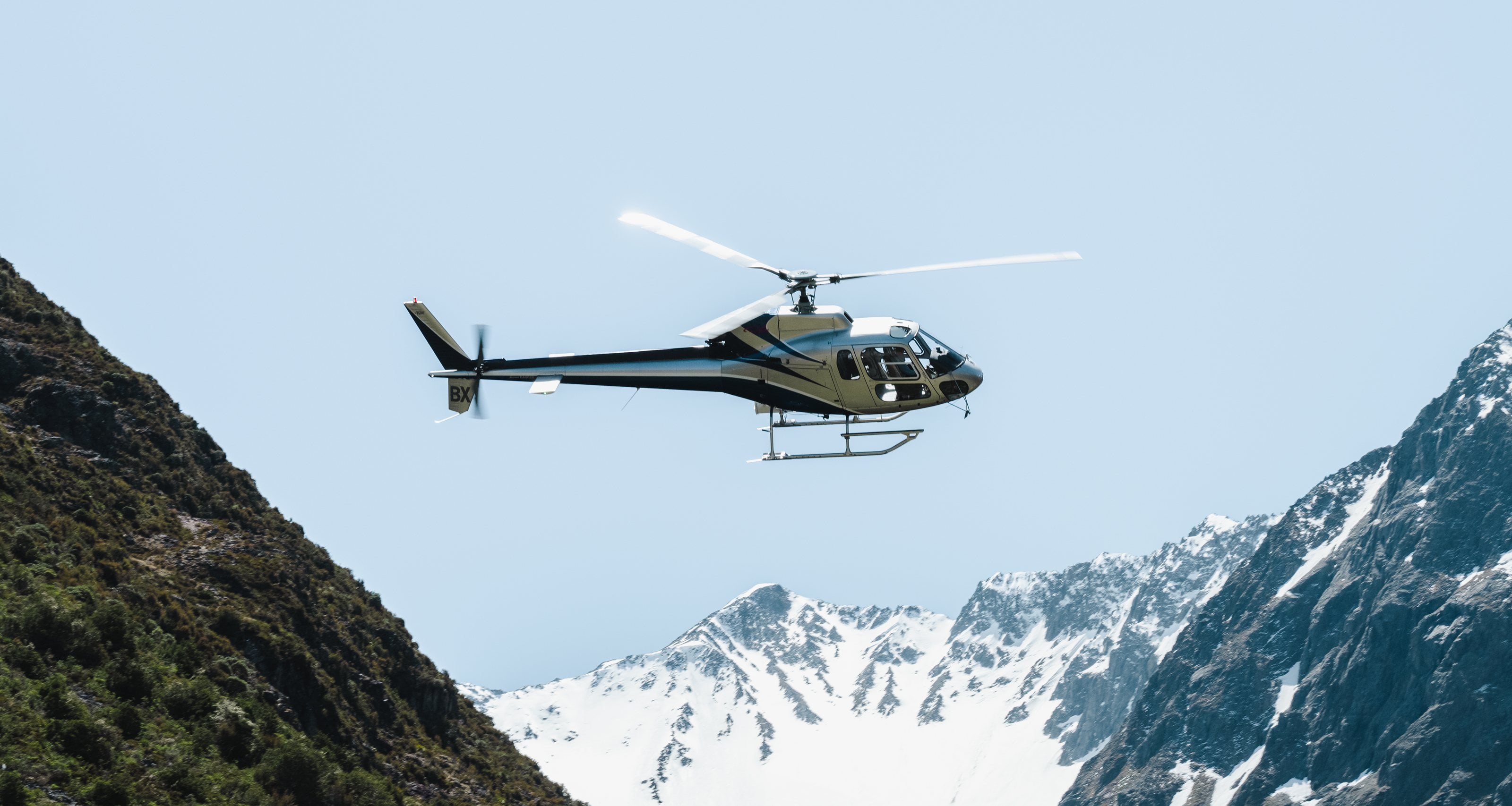 A helicopter in flight between bush and snow-covered peaks in the Southern Alps