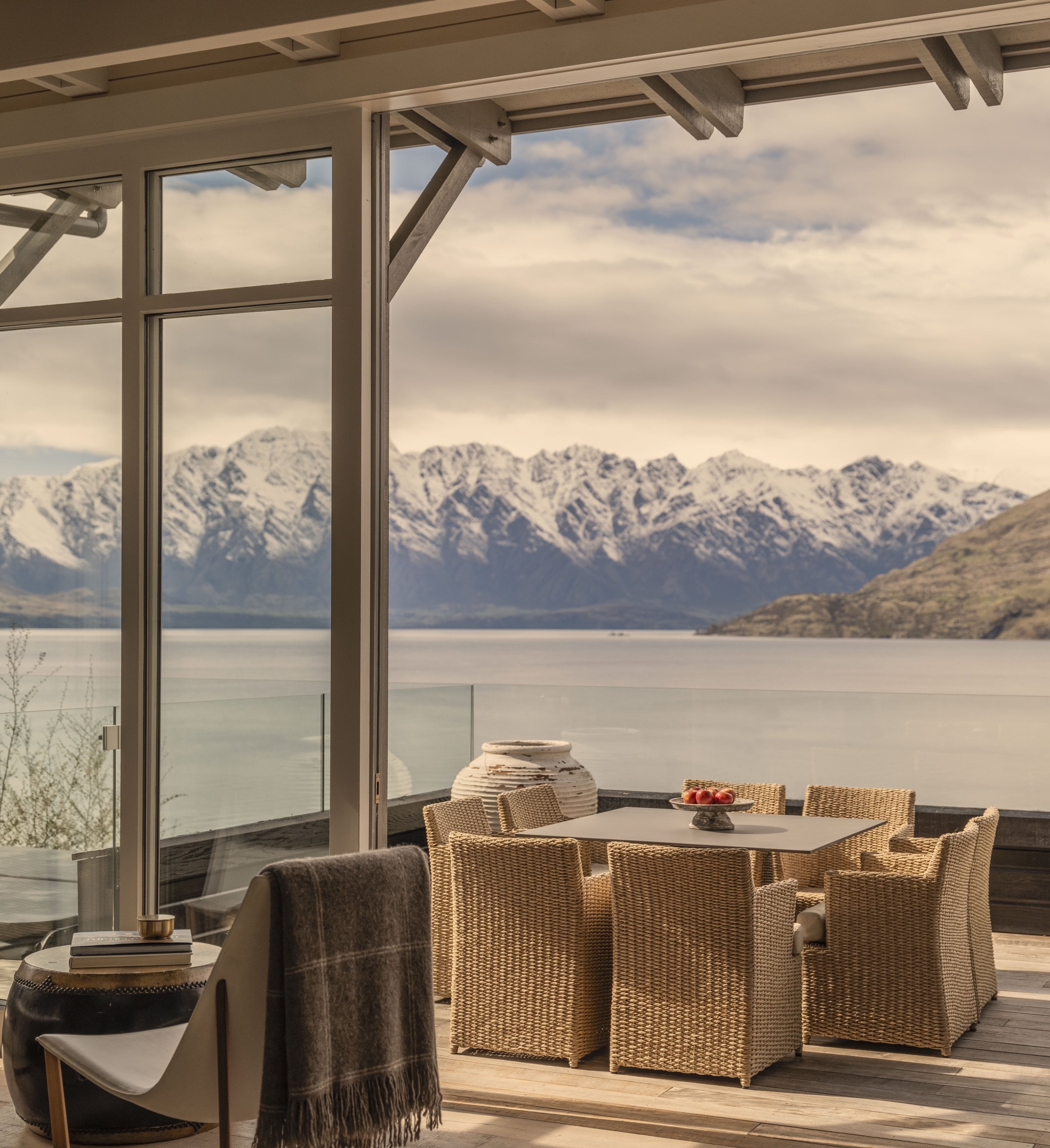 A terrace at Rosewood Matakauri, wicker chairs set before Lake Wakatipu and the snow-covered Remarkables