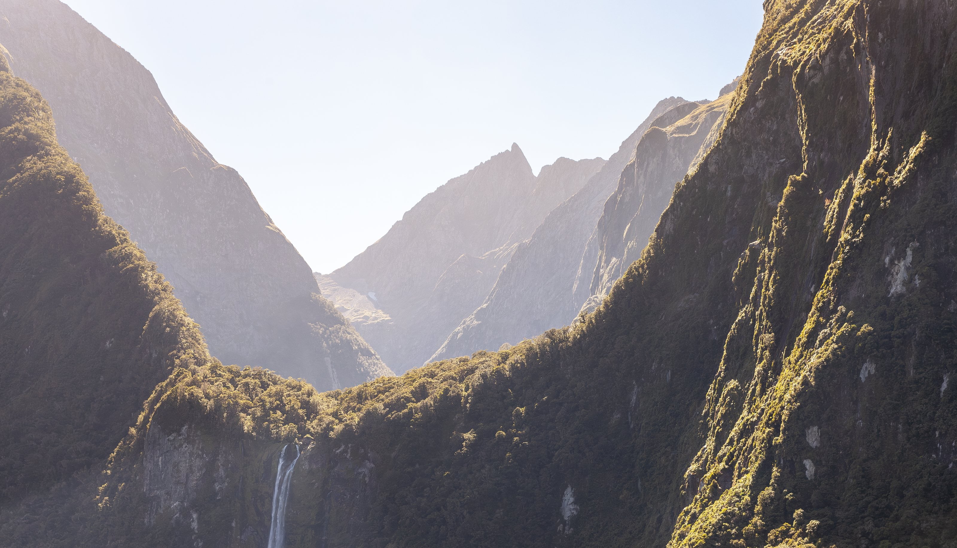 A waterfall off a bush-covered cliff in a fiord, Fiordland, the peaks in haze