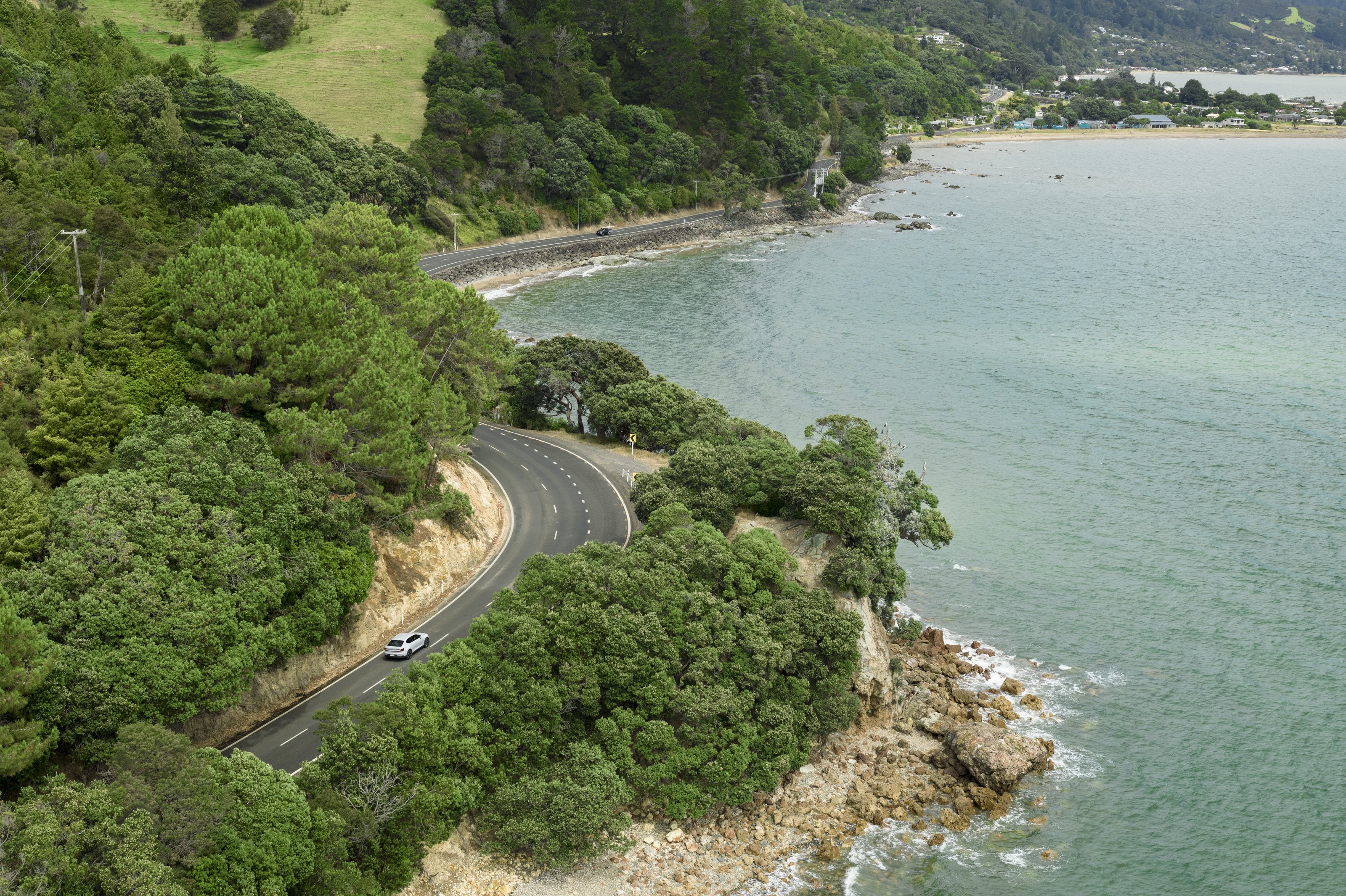 A car on a coastal road curving above a bay, seen from the air