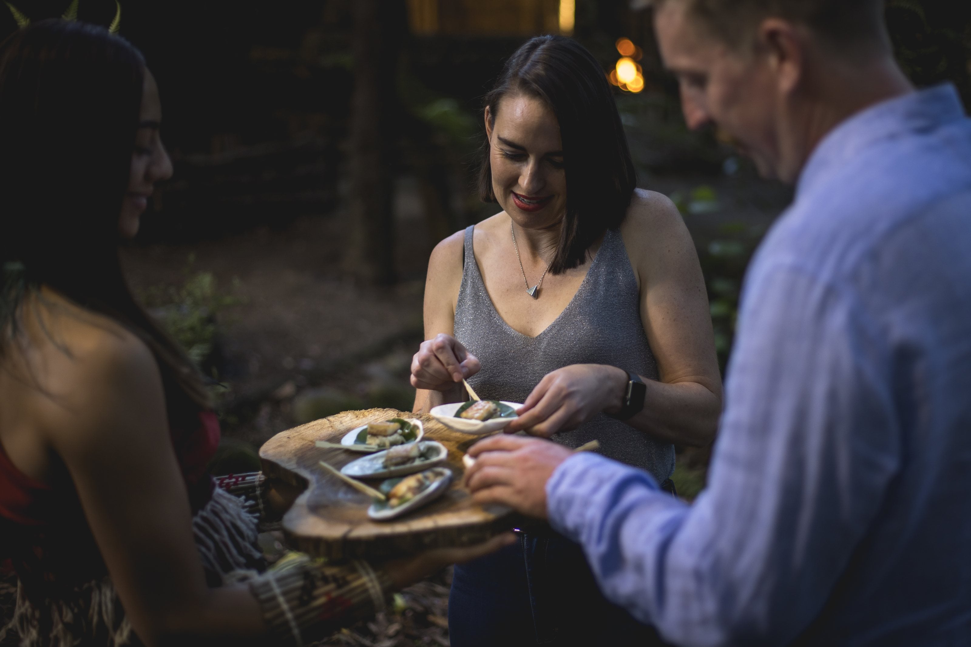 Three guests sharing small plates from a wooden board outdoors at dusk
