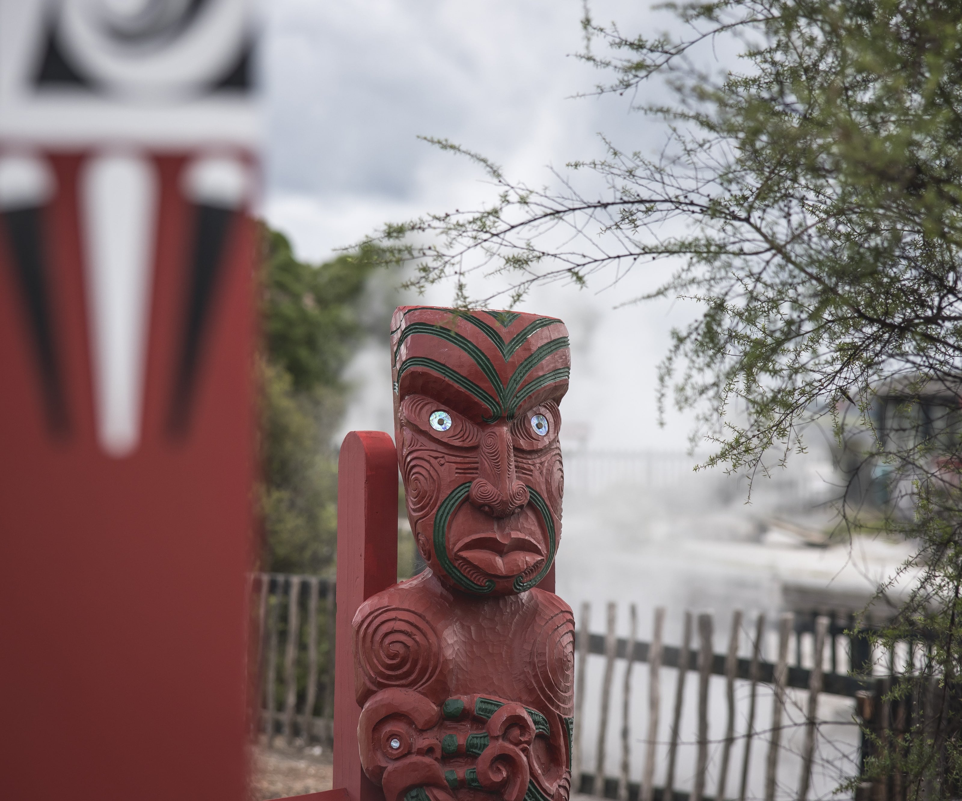 A carved red pou with pāua-shell eyes, geothermal steam rising behind it, Rotorua