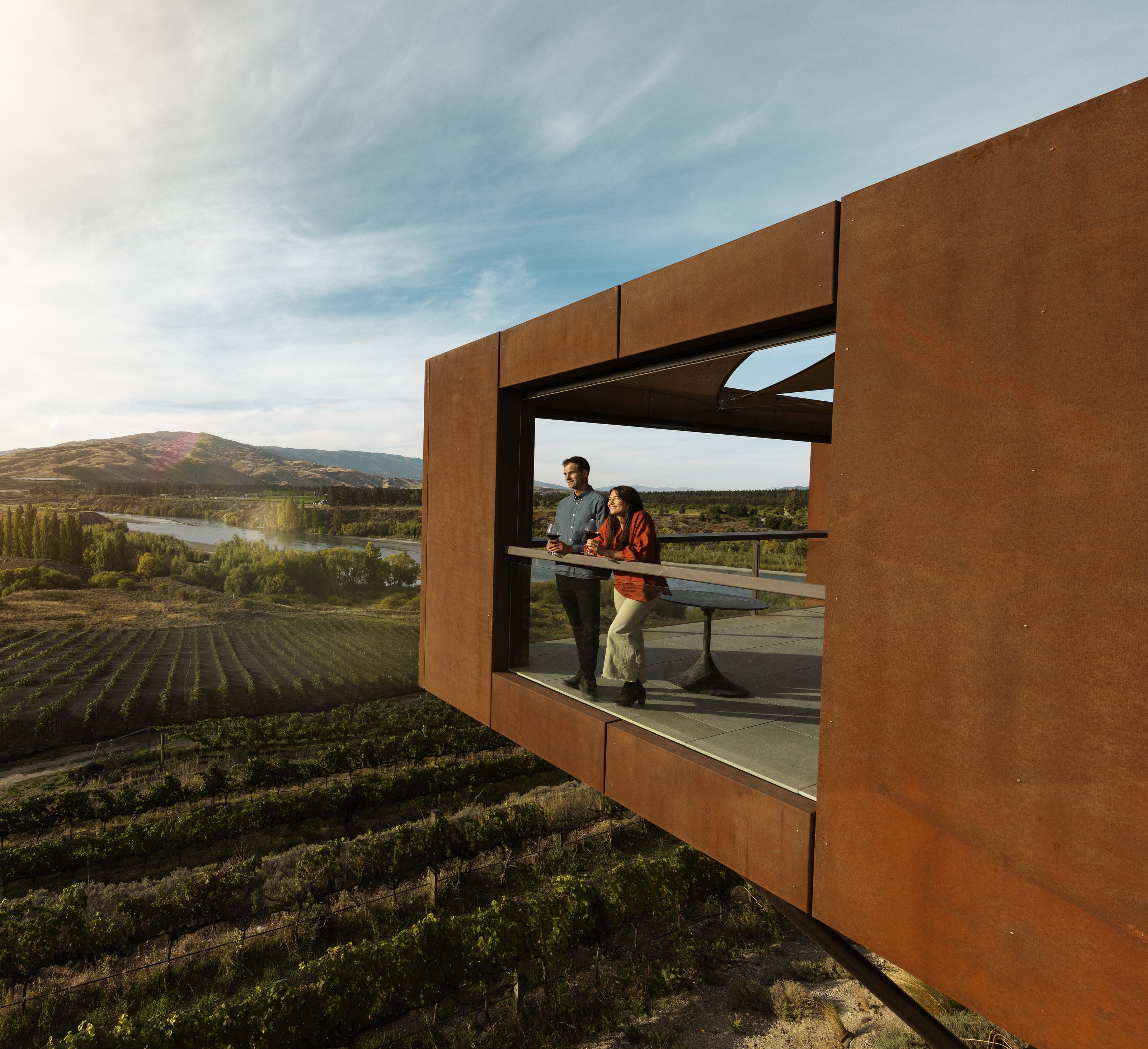 Two guests with glasses of red on a rusted-steel lookout above the vineyards, Central Otago
