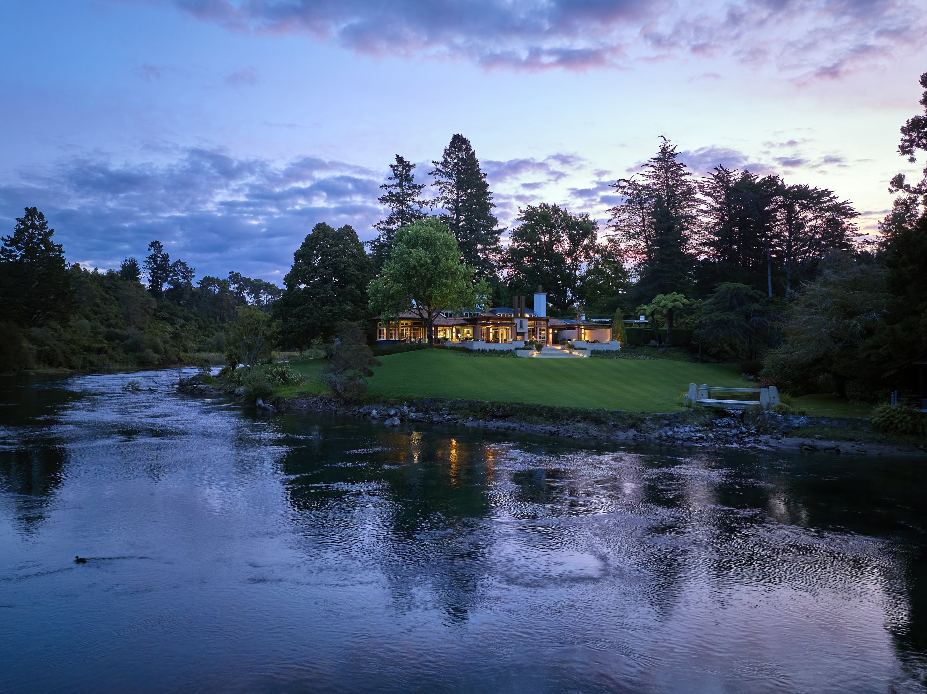 Huka Lodge lit at dusk across the Waikato River, its lawn running to the water