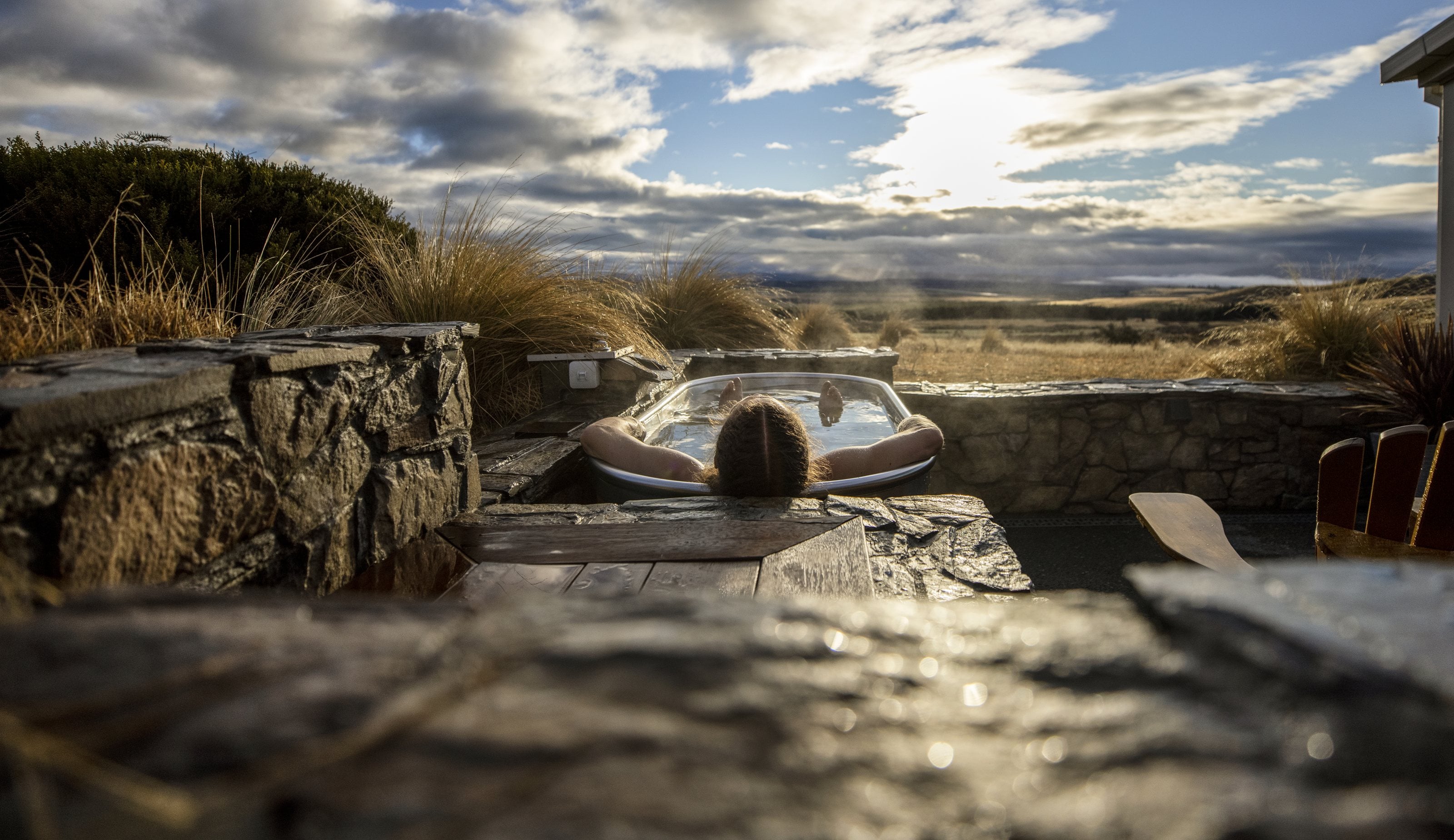 A guest in an outdoor bath within a stone-walled courtyard, tussock and the Mackenzie plain beyond