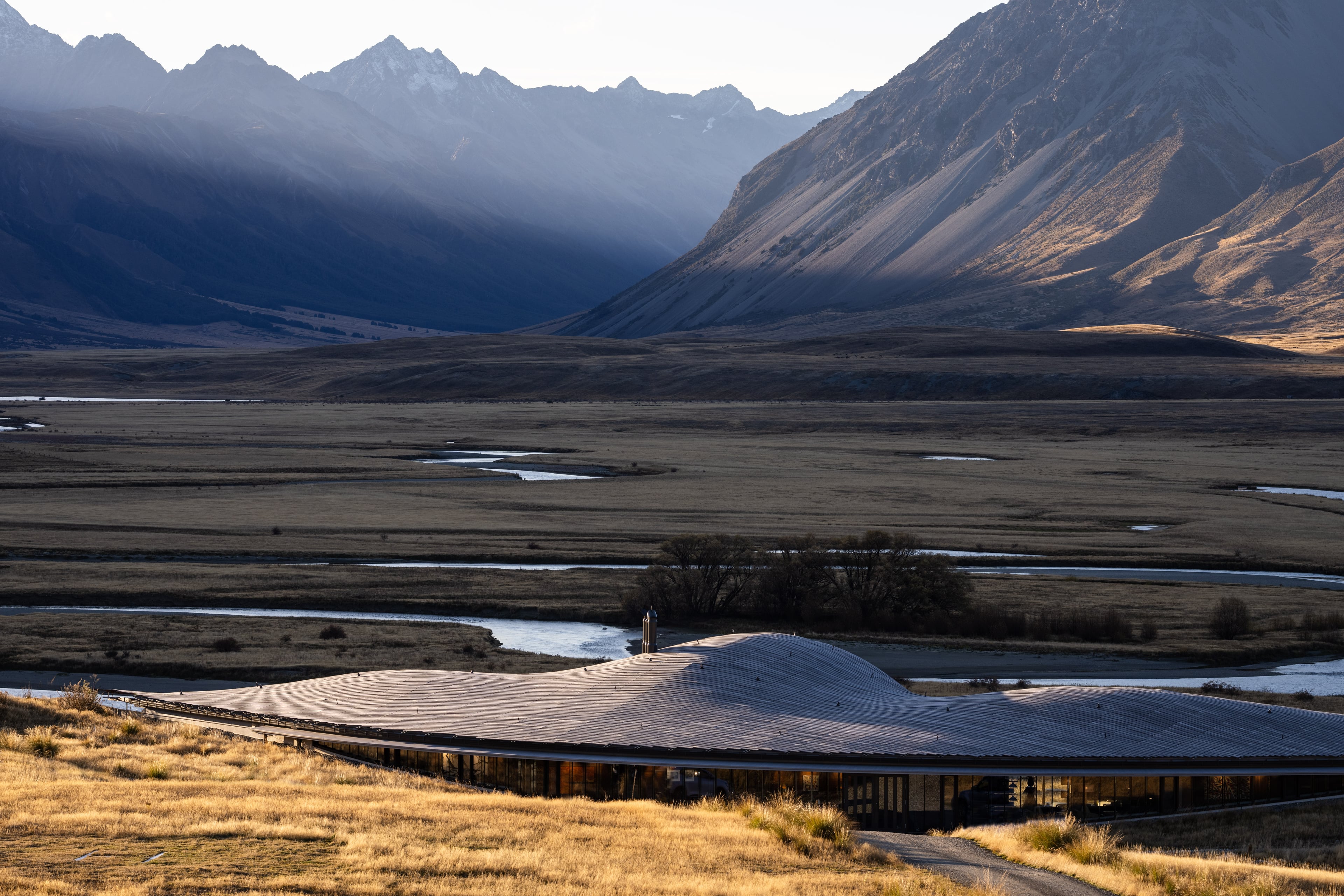 The long curved roof of The Lindis low in golden tussock above the braided Ahuriri River, the valley and mountains beyond in late light